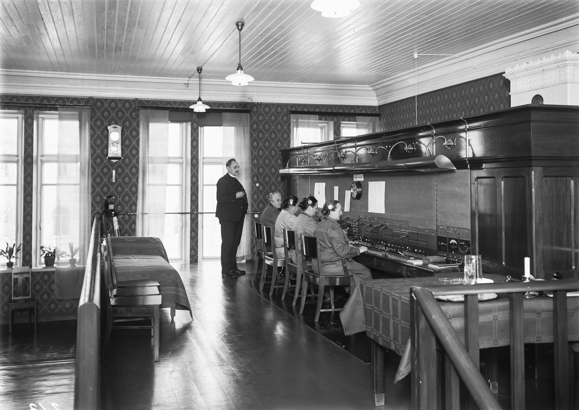 Operators seated along a long manual switchboard in a panelled exchange hall, a supervisor standing behind them